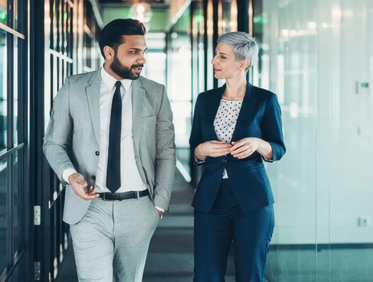 A man in a light gray suit and a woman in a navy suit walk together in a modern office hallway, engaged in conversation. Both look professional and focused.