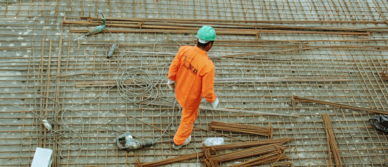 A construction worker in an orange jumpsuit and green helmet stands on a metal grid with scattered rebar and tools at a building site, seen from above.
