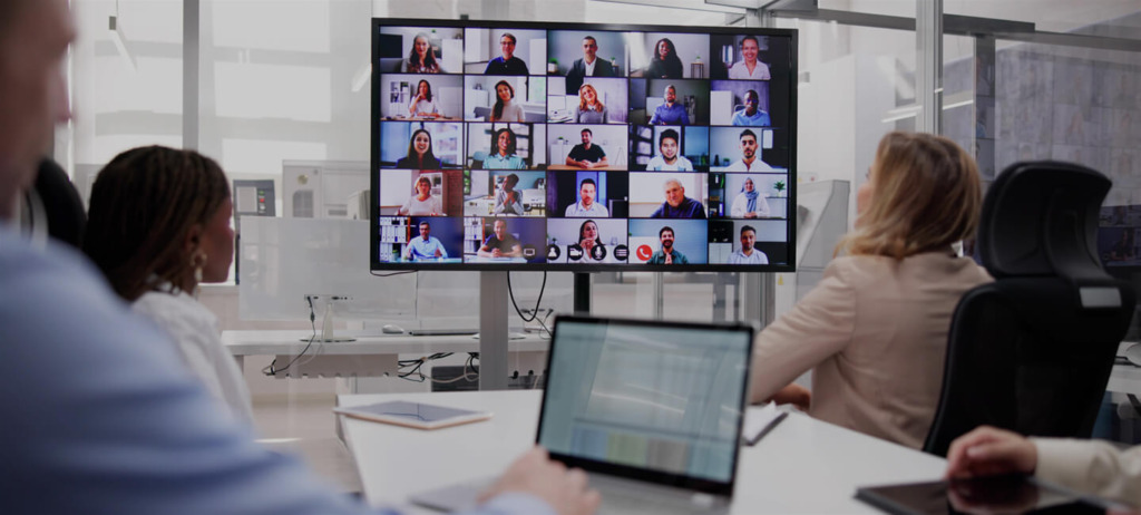 People in an office conference room attend a virtual meeting with many participants visible on a large screen.