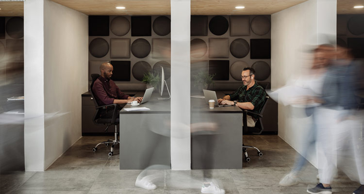 Two men work at computers at separate desks in a modern office with geometric wall decor. Several people walk by in front of them, appearing blurred due to movement.