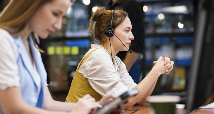 Two women working at desks in an office, one wearing a headset and looking at a computer screen, the other using a tablet. The background is blurred with lights and shelves visible.