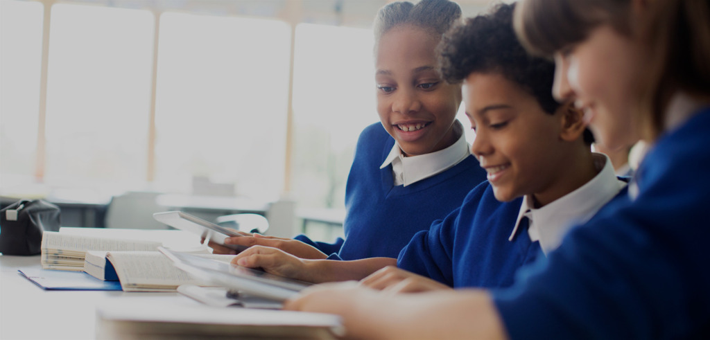 Three students in blue school uniforms sit at a table, smiling and looking at a tablet together, with open books nearby in a bright classroom setting.