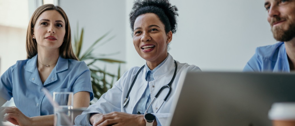 Three medical professionals, two women and one man, sit together at a table in a bright room. One woman wears a stethoscope and smiles while listening, suggesting a collaborative meeting or discussion.