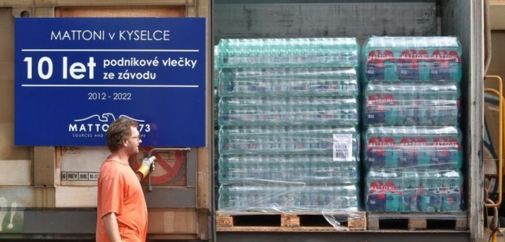 A man in an orange shirt stands near a truck loaded with stacked pallets of bottled water. A blue sign in Czech celebrates ten years of business from 2012 to 2022 at the Mattoni factory in Kyselka.