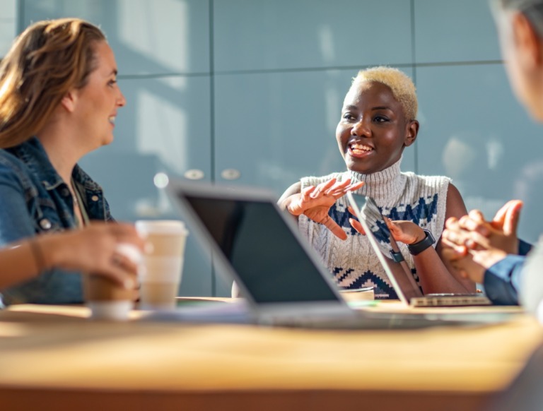 Three people sit at a table with laptops and coffee cups, engaged in a lively discussion. One person gestures while speaking, and the group appears to be collaborating in a bright, modern office setting.